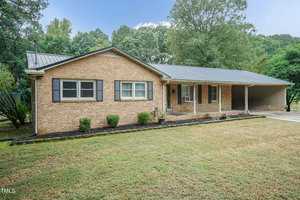 A single-story brick house with a metal roof, featuring a covered front porch supported by four columns and a carport to the right. The yard is partially landscaped with shrubs and trees in the background. The sky is clear, and the overall setting is calm and suburban.