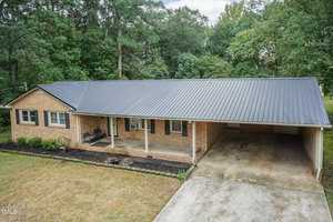 A single-story brick house with a metal roof, featuring a front porch with seating, a neat garden bed running along the front, and an attached carport on the right. The background shows tall trees surrounding the property. The driveway leads up to the carport area.