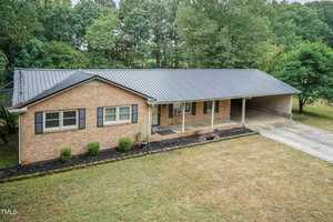 A single-story brick house with a large covered carport and a metal roof. The front yard has a mix of grass and small shrubs along the foundation. The background features tall, dense trees. The overall setting is suburban and peaceful.