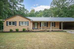 A single-story brick house at 2788 Vicksboro Road with a covered front porch, a glass-paneled front door, and three front-facing windows. The home is surrounded by a neatly mowed lawn, small shrubs, and is situated next to a wooded area in Henderson. An attached carport is on the right side.