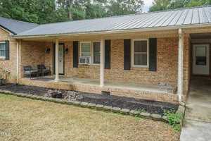 A single-story brick house with a metal roof features a covered front porch. There are two wicker chairs and a small table on the porch. Black window shutters and a window air conditioning unit are visible. The lawn is neatly trimmed, and there are garden beds along the porch.