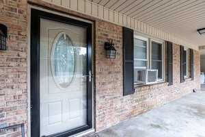 A residential brick house features a covered porch with a concrete floor. The front door is white with a decorative oval glass insert framed in black. Beside the door, there is a wall-mounted black lantern. Two windows with black shutters have window-mounted air conditioners.