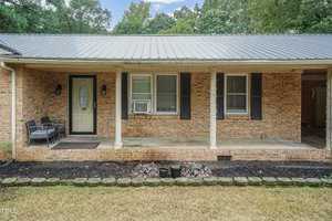 Brick house with a covered front porch. The porch has a black front door with an oval window, two black chairs, and string lights along the columns. Three windows with black shutters are visible. The yard has a narrow stone border and some small plants. Trees are in the background.