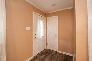 A small entryway with tan walls, a door with a glass window covered by a sheer curtain, and another closed white door. The floor is covered in wood-patterned vinyl, and the ceiling has crown molding. A light switch and vent cover are visible on the left wall.