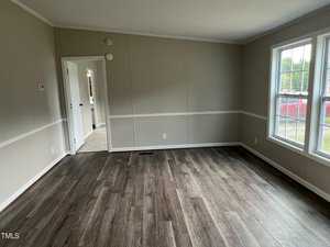 A bright, empty room with light beige walls, white trim, and wood-look laminate flooring. Large windows on the right allow natural light to enter. An open doorway on the left leads to another room, and another door is visible in the background.