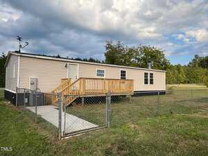 A single-story beige mobile home with a wooden front porch sits on a grassy lawn, surrounded by a chain-link fence. The home is set against a backdrop of trees and a partly cloudy sky. An antenna is visible on the roof.