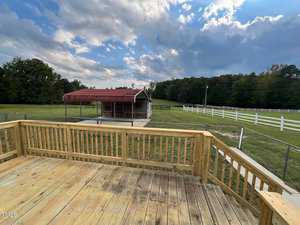 A wooden deck overlooks a spacious, grassy yard with a small red-roofed shed. The area is fenced with white fencing, and the background features trees and a partly cloudy sky.
