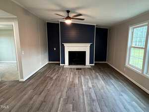 A spacious living room with laminate wood flooring, beige walls, and a dark blue accent wall behind a white fireplace. A ceiling fan with a light is mounted in the center of the ceiling, and two large windows allow natural light into the room. A doorway leads to an adjacent carpeted room.