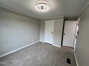 A small, empty room with beige carpet, light gray walls, and a white ceiling. It has a round ceiling light fixture, a closed white door, and an ajar door leading to another space. A small floor vent is visible in the corner.