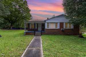 Located at 804 Eastside Drive, this charming single-story brick house in Henderson features a welcoming porch and steps leading to the front door. The yard boasts lush grass, trees, and a concrete path. The sky is alive with pink and orange hues, signaling a serene sunset or sunrise.