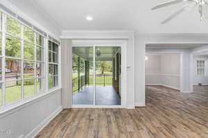 A bright, open room with large windows on the left, sliding glass doors leading to an outdoor area in the center, and an open doorway to another room on the right. The room has light-colored walls, wood flooring, and a ceiling fan.