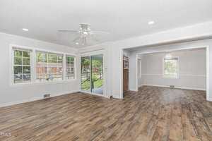 Spacious living area with light wood flooring, ceiling fan, large windows, and sliding doors leading to a porch. Adjacent room with similar flooring and a chandelier. White walls and molding add a modern, airy feel.