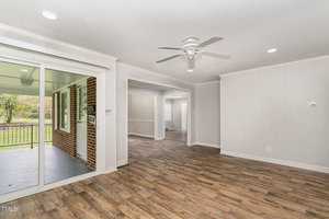 A bright, empty living room with light-colored walls, recessed lighting, and a ceiling fan. The room features wood flooring and a large sliding glass door leading to a enclosed porch with brick walls and windows overlooking a green, outdoor area.