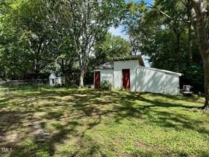 A small white shed with a red door stands in a shaded backyard surrounded by trees. A shorter adjacent extension with a similar appearance is attached to the shed. The ground is covered with grass and patches of dirt, and a chain-link fence is visible on the left.