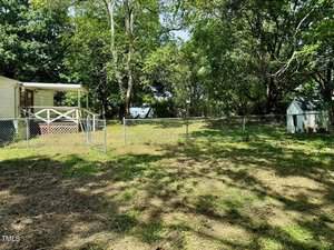 A fenced backyard with a grassy area and trees providing shade. There is a small white shed on the right side and a white house with a covered porch on the left. The background shows a view of the woods, giving the yard a secluded and peaceful atmosphere.