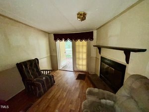 A modest living room with a dark wooden rocking chair and a cushioned armchair facing a fireplace. Light filters through glass doors covered by blinds and a valance, leading to an outdoor deck. The room has a wooden floor and plain, light-colored walls.