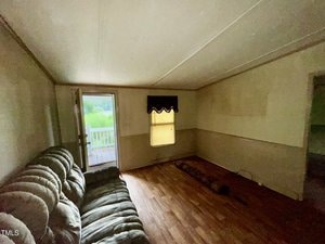 A dimly lit room with light-colored walls, a wooden floor, and a green, fabric sectional sofa. An open door leads to a deck outside. A window with a black valance is centered on the back wall, allowing some natural light in. The room is mostly empty and appears to be in need of some finishing.