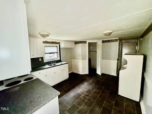 A kitchen with white cabinets, dark countertops, and a small sink under a window. The room has dark tile flooring and a stove on the left. There is a small open closet and a doorway to the right. The walls are light-colored with a decorative border.