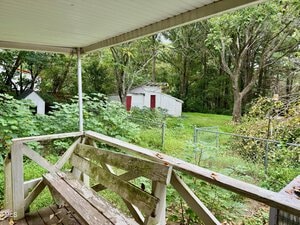 A backyard with a covered wooden porch featuring a weathered bench. The yard is lush with vegetation and trees, and a white storage shed with red doors is visible in the distance. A metal fence runs along the edge of the property.