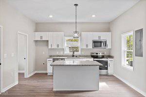 A modern kitchen with white cabinets, a center island with granite countertops, stainless steel appliances, and wooden flooring. Large windows provide natural light, and a pendant light hangs above the island. Walls are painted light gray.