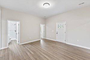 A bright, empty room with light wood flooring and neutral walls. It features a ceiling light, a door with a window showing a stained glass pattern, and an open door leading to a tiled bathroom.