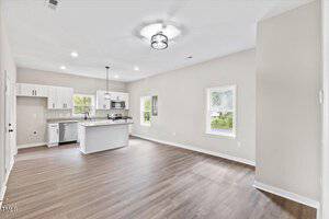 Bright, modern kitchen and living area with white cabinets, an island, stainless steel appliances, and wood flooring. Two large windows provide natural light, under a ceiling light fixture. Walls are painted light gray.