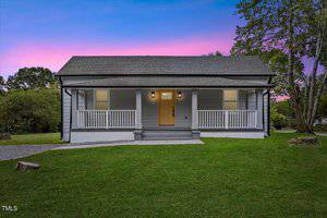 A small, single-story house on 916 Eaton Street in Henderson boasts a wooden front porch with white railings against a sunset sky. The neatly trimmed lawn and surrounding trees enhance its charm. A wooden front door and two lamps warmly illuminate the inviting porch.