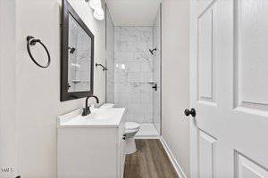 A small, modern bathroom with white walls and a marble-tiled shower. It features a white vanity with a black-framed mirror, black fixtures, and a wooden floor. The design is minimalistic and elegant.