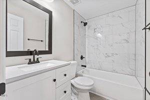 A modern bathroom with white marble tiles in the shower, a white vanity with a black-framed mirror, black fixtures, and a white toilet. The shower features a dark showerhead and faucet. Walls are painted light gray.