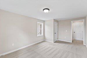 A bright, empty room with beige walls and clean light beige carpet. There is a window letting in natural light, and two white doors leading to adjacent areas. A ceiling light fixture is centered above.