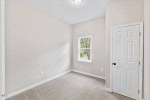 An empty room with light gray walls, a white door on the right, and a window showing a tree outside. The floor is carpeted in a neutral shade, and the room is well-lit with natural light.