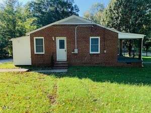 A small brick house with a white front door and two windows. It has a grassy front yard with scattered fallen leaves. There is a small porch on the right side and trees in the background under a clear blue sky.