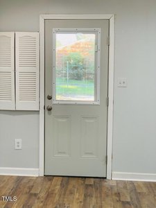 A light gray door with a rectangular window near the top, set in a white frame. The door is in a room with light gray walls and a wooden floor. There are louvered panels on the left wall and green grass visible through the window.