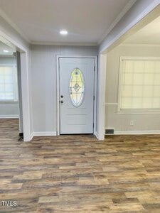 A bright room with light-colored walls and wood laminate flooring. The focus is on a closed white door with an oval, stained glass window at its center. The space is well-lit, with natural light streaming through a window with blinds.