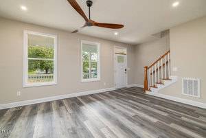 A spacious, unfurnished living room with light gray walls and wood-patterned flooring. The room features two windows, a ceiling fan, and a staircase to the right. A front door with a small window is visible in the background.