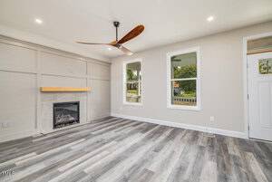 A modern living room with light wood flooring and a recessed ceiling light. There is a fireplace with a wooden mantel on the left and two windows on the right. A ceiling fan with wooden blades hangs in the center of the room.