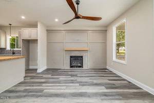 A modern living room with light gray walls and wood-look flooring. The room features a central fireplace with a wooden mantel and a ceiling fan with wooden blades. A window on the right lets in natural light, and the kitchen is partially visible on the left.