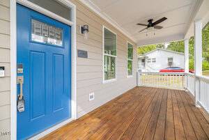 A charming porch with a blue door, wood flooring, and light beige siding. A ceiling fan is mounted above. Large windows allow natural light, and trees and a neighboring house are visible in the background.
