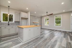 Modern kitchen with light gray cabinets and a wood-topped island. Open layout features wood-look flooring, a ceiling fan, large windows, and recessed lighting. White walls enhance the brightness of the space.