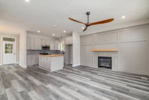 A modern, open-concept living area with light wood flooring. It features a sleek fireplace, ceiling fan, and a kitchen with gray cabinetry, a central island, and stainless steel appliances. A door and windows provide natural light.