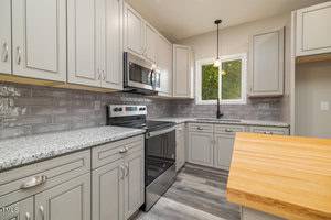 A modern kitchen with light gray cabinets, stainless steel appliances, and a granite countertop. There's a wooden island in the foreground, a pendant light above, and a window providing natural light. The backsplash features a glossy subway tile design.