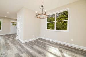 A bright, empty room with light wood laminate flooring, beige walls, and a large window showing green foliage. A black metal chandelier with exposed bulbs hangs from the ceiling, and a doorway leads to another room.