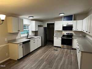 A spacious, modern kitchen with white cabinets, gray countertops, and stainless steel appliances. The room features a large window above the sink, wood-style flooring, and recessed lighting, creating a bright and inviting atmosphere.