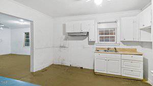 A clean, empty kitchen with white cabinets, a sink under a window, and a range hood. The walls and ceiling are white, and the floor appears to be unfinished or covered with brown protective material. There's an open doorway to another room.