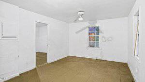 A small, empty room with white walls and a tan floor. There is a ceiling fan, one window, and a doorway. The walls show signs of wear and patches of discoloration. Natural light filters through the window.