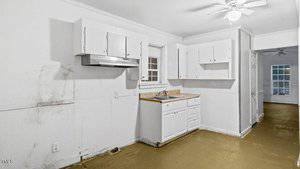 A small, empty kitchen with white cabinets and a sink. The walls are white with visible stains, and the floor is bare. A ceiling fan with a light is on the ceiling, and there is a window above the sink.