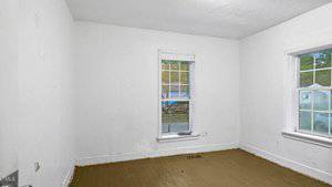 A small empty room with white walls and a wooden floor. Two windows with white trim let in natural light. The paint is slightly peeling near the baseboard and the floor has some marks. Outside, greenery and a slightly visible car are seen through the windows.