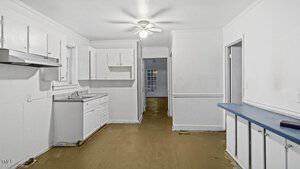 A small, empty, white-walled kitchen with a sink and cabinets on the left. A narrow hallway extends to the back, leading to a room with a window. A ceiling fan is above, and there's a narrow counter on the right side. The floor is a plain, brown color.