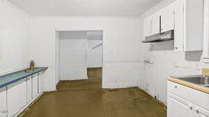 A minimalist kitchen with white walls and cabinets. The floor appears to be unfinished. A single sink and range hood are visible. Through an open doorway, another empty room can be seen. A small decorative item sits on a blue counter.