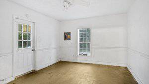 An empty room with plain white walls and a bare floor. There is a window to the right and a door with a window on the left. A ceiling fixture is visible, and the room has minimal decor and no furniture.